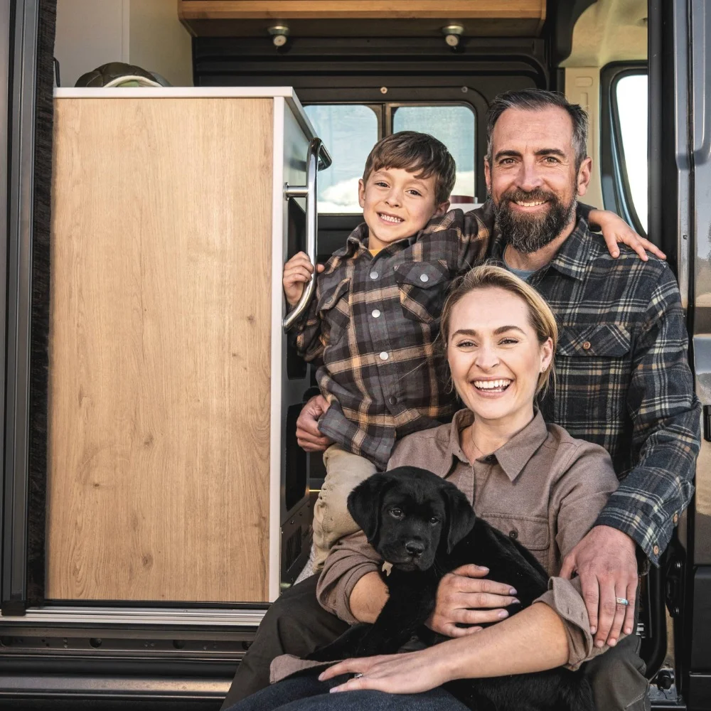 Smiling family with a young boy and a black puppy poses inside a van, showcasing a cozy and cheerful atmosphere.