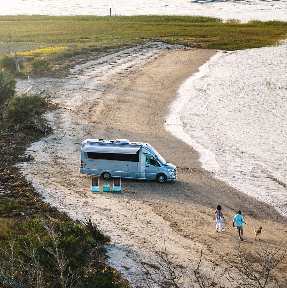 Aerial view of a camper van parked on a sandy beach near the water, with two people walking and a picnic setup nearby.