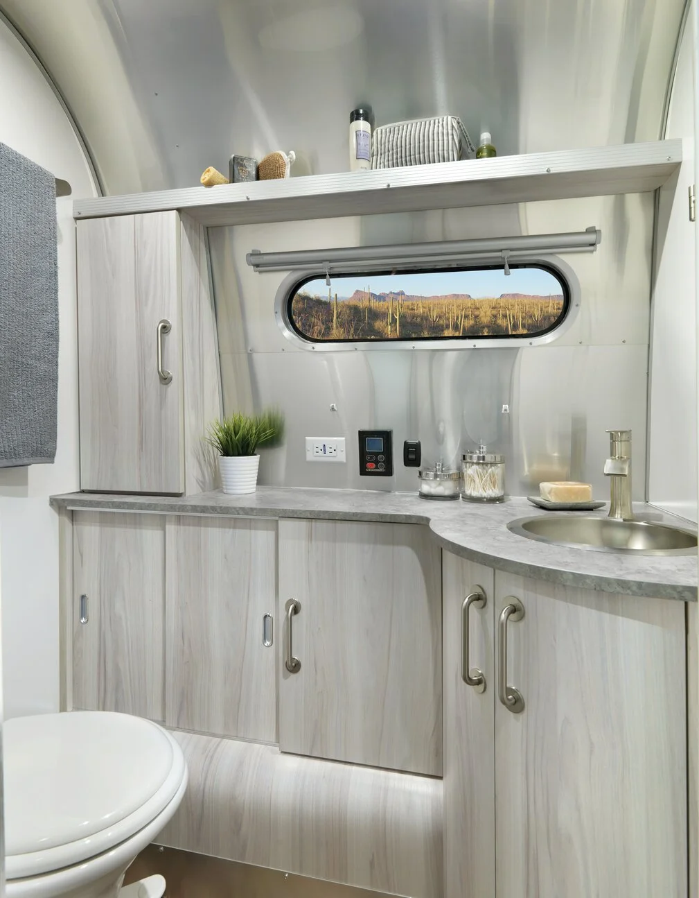 Modern RV bathroom with light wood cabinets, oval window view of cacti, a round sink, and a toilet. Towel and toiletries on shelves.