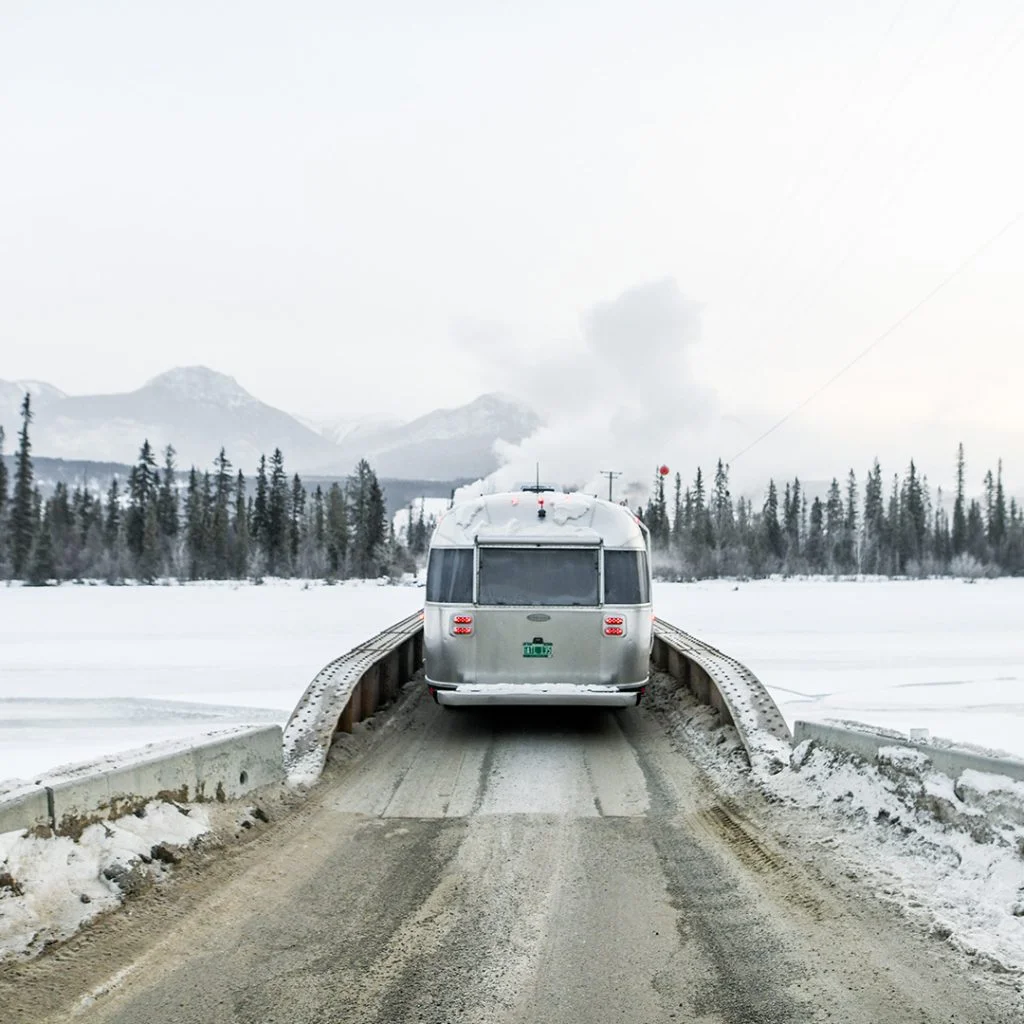 RV driving on snowy bridge surrounded by frozen landscape and mountains, under an overcast sky.