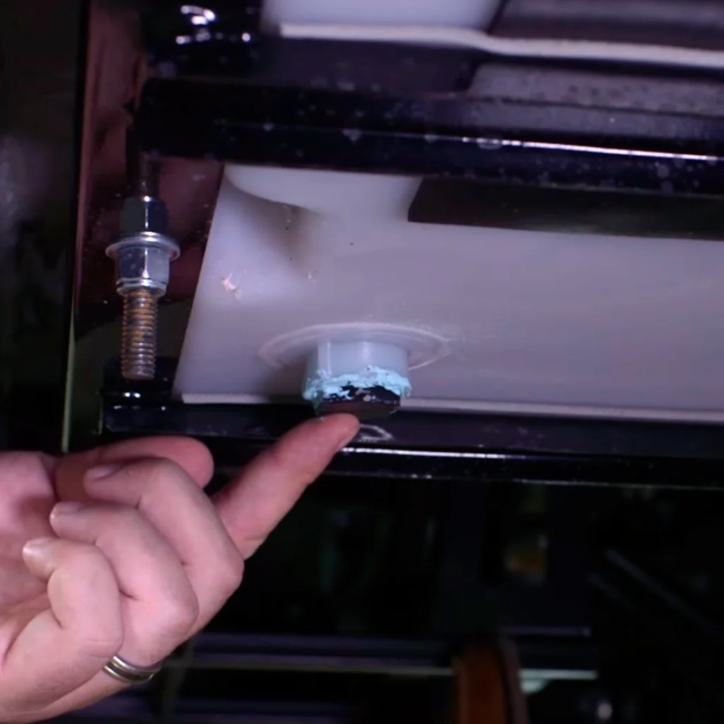Hand pointing at a blue, corroded bolt under a white plastic tank, with surrounding black metal framework.
