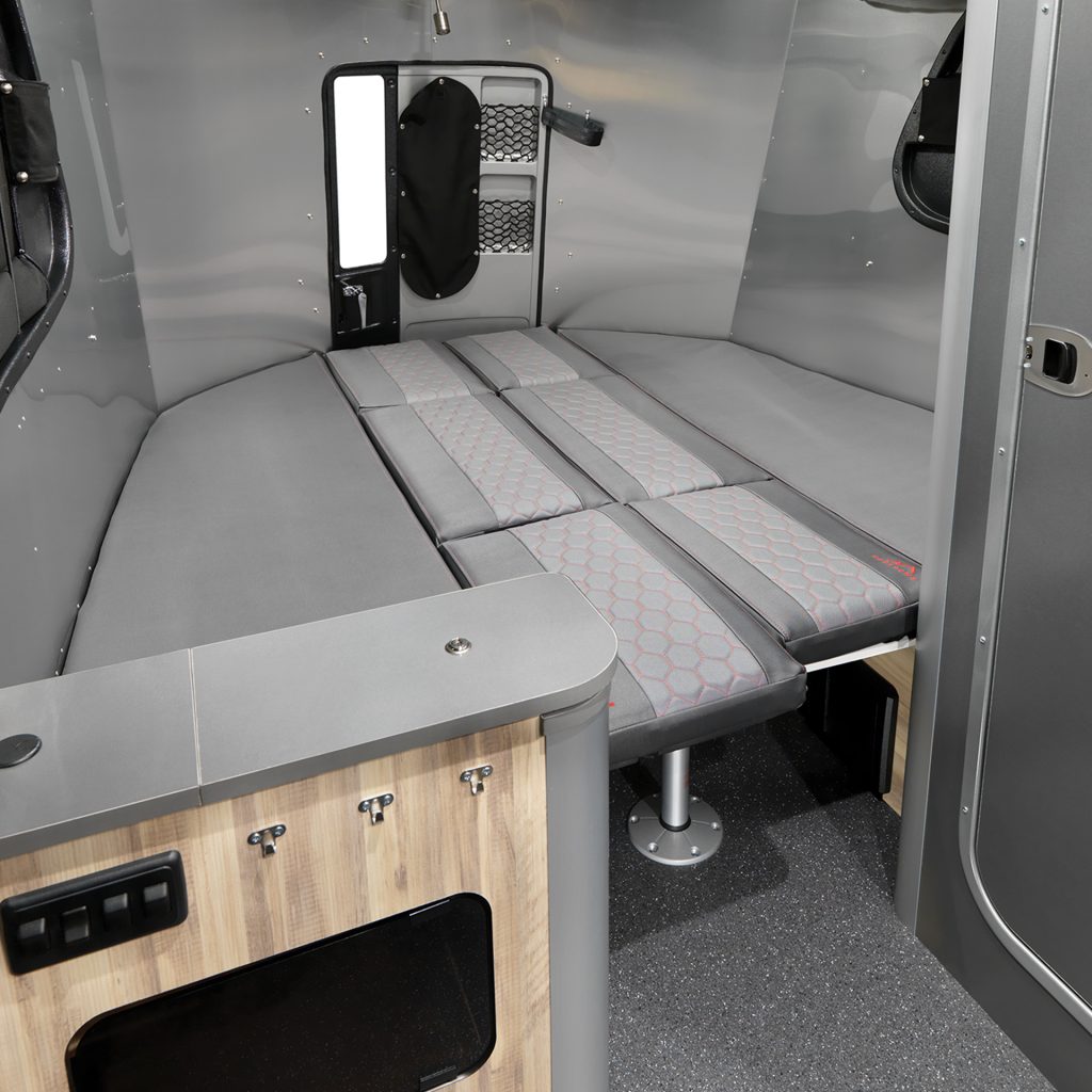 Compact camper interior with grey padded seating and a fold-out bed, wooden storage cabinet, small window, and modern design elements.