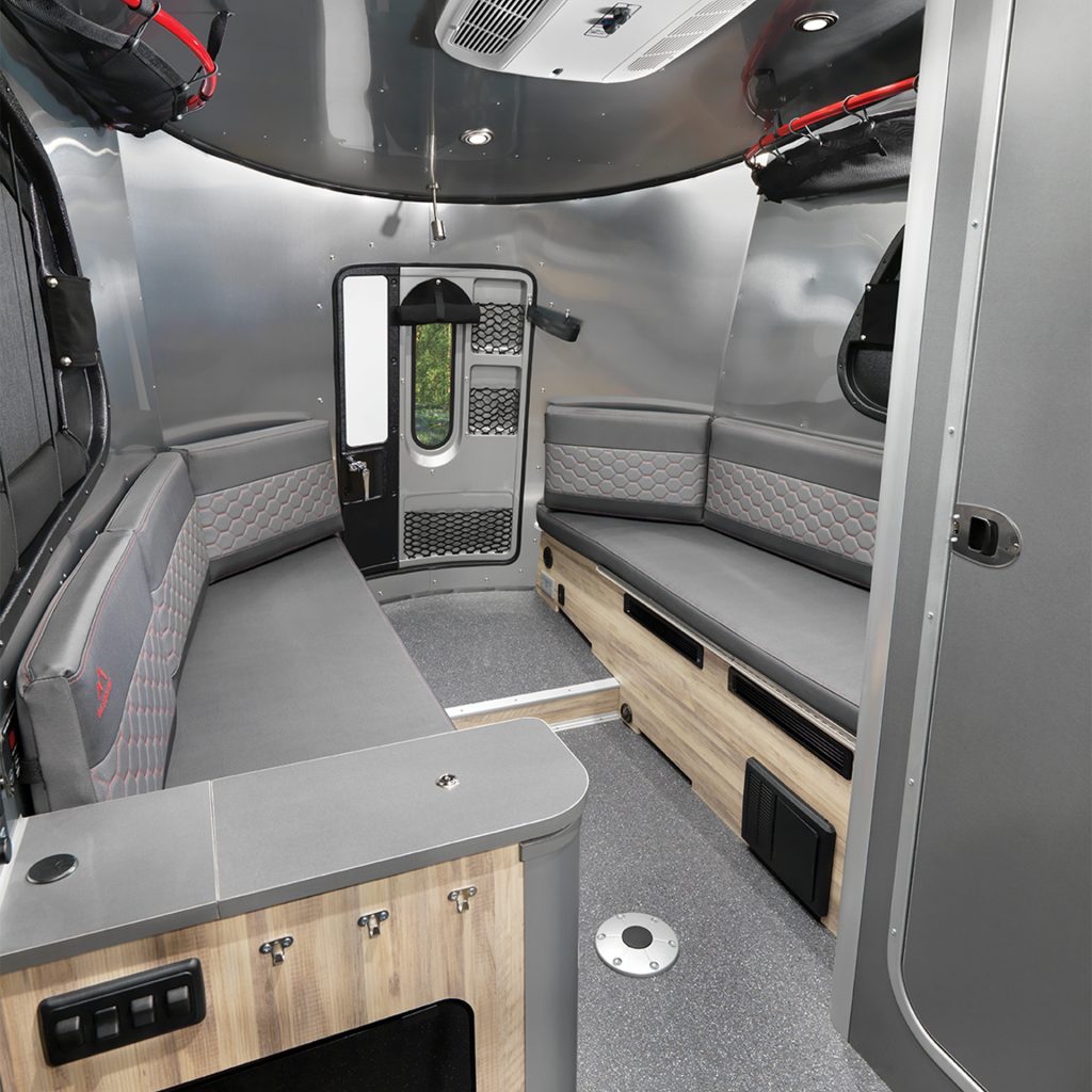 Modern RV interior with sleek gray sofas, wood accents, and a central door. The ceiling has built-in lighting and an air vent.