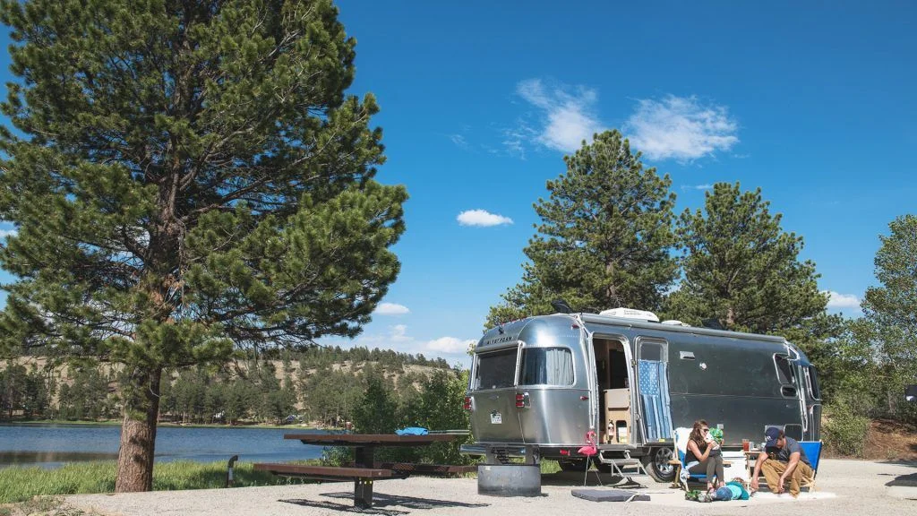A silver camper trailer is parked at a lakeside campsite surrounded by pine trees. People are sitting on a picnic table and relaxing on a blanket nearby under a blue sky with scattered clouds.