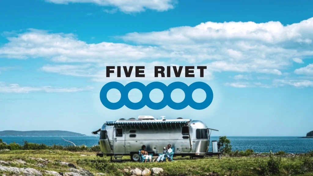 A silver RV is parked by the sea with two people seated outside. Blue sky and scattered clouds in the background. Text reads "FIVE RIVET."