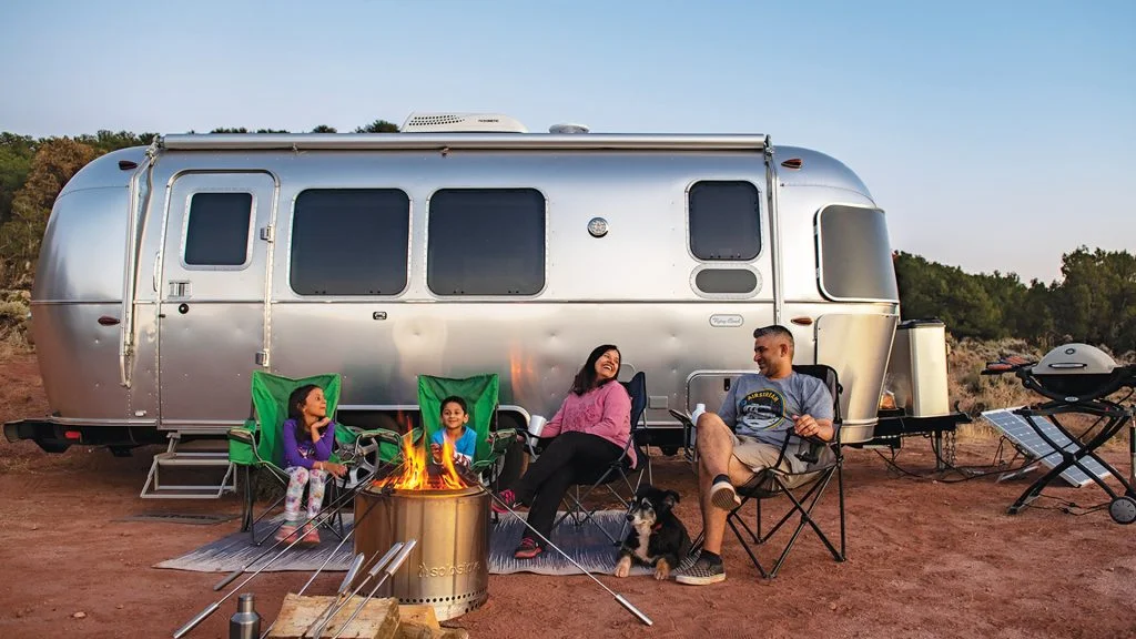 Family camping by a silver trailer, sitting around a campfire with green chairs and a dog. Barbecue and solar panel nearby.