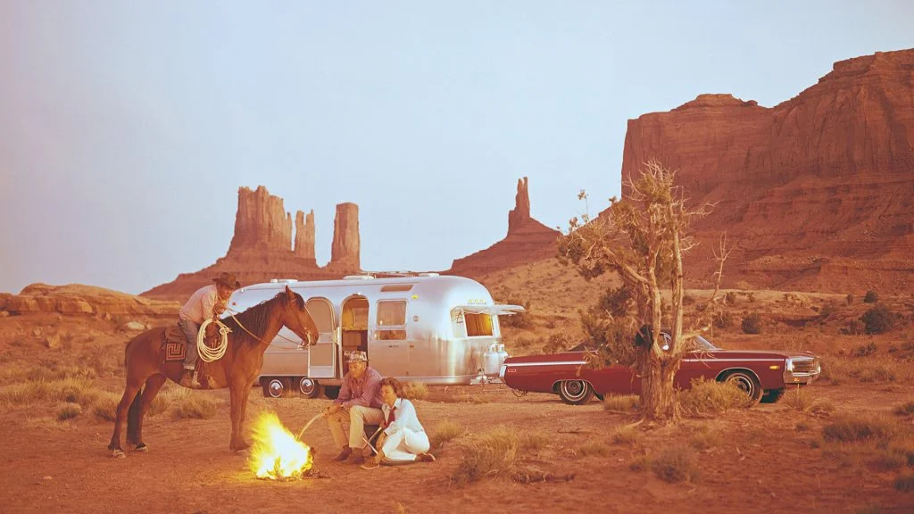 Three people and a horse are in a desert setting by a campfire. Behind them are a silver Airstream trailer and a red classic car, with tall rock formations visible in the background.