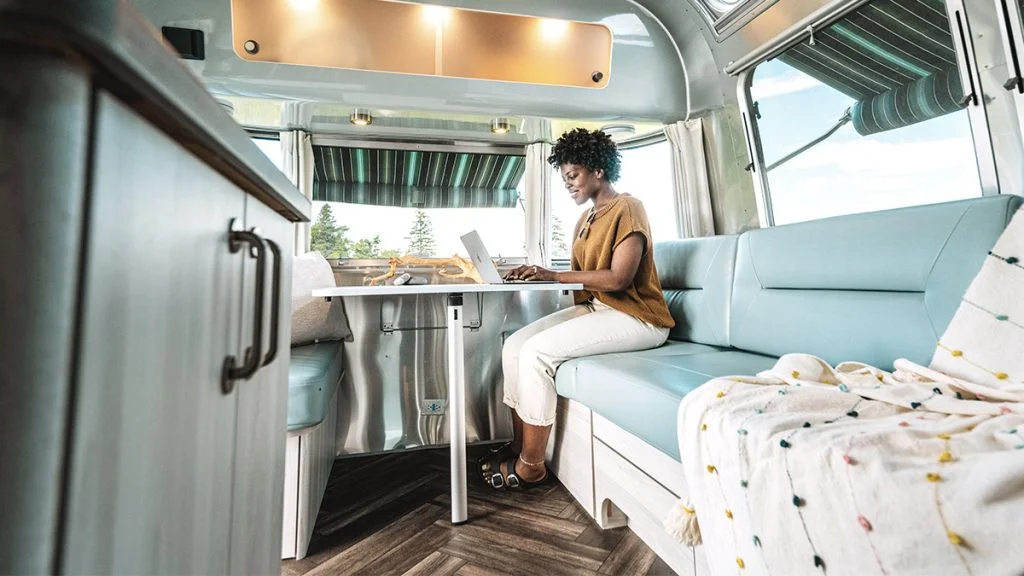 A woman works on a laptop at a table inside a modern RV with light blue seating, wood flooring, and large windows showing a sunny outdoor scene. A patterned blanket is draped over the couch.