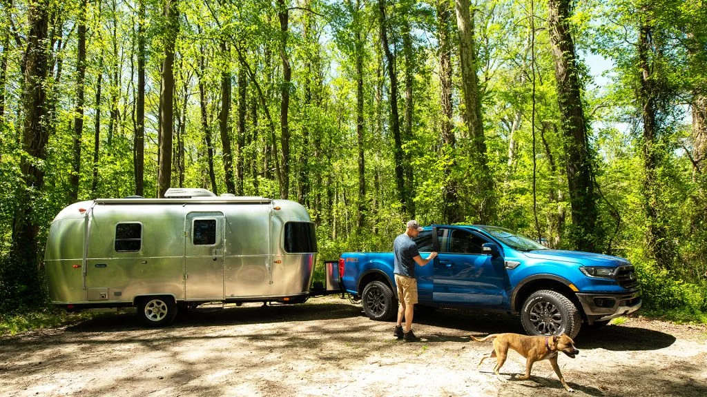 A man stands next to a blue truck hitched to a silver trailer in a sunlit forest, while a brown dog walks nearby.