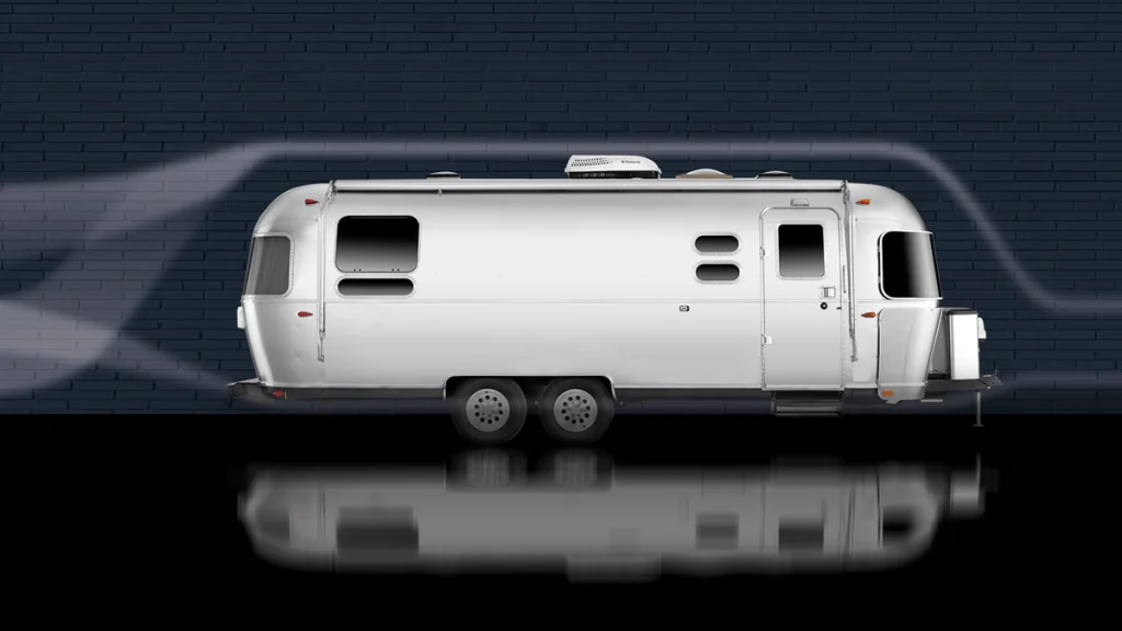 A silver Airstream travel trailer is parked in front of a dark brick wall, with airflow lines illustrating aerodynamics over its smooth, rounded surface. The trailer’s reflection is visible on the glossy ground.