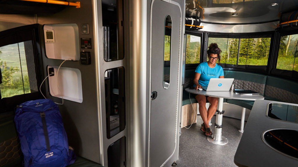 Person working on a laptop in a modern RV with curved windows, surrounded by trees. A blue backpack is visible nearby.