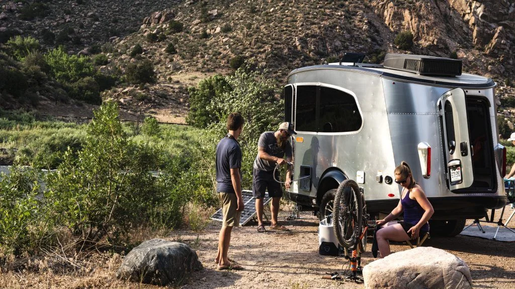 Three people set up camp beside a silver trailer in a rocky, mountainous area. One person works on a bicycle, another stands nearby, and the third attends to the trailer. Green bushes and rocks surround the campsite.