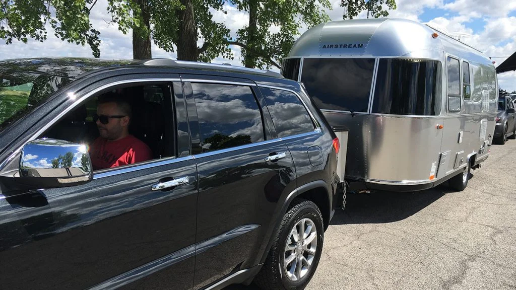 A man drives a black SUV towing a silver Airstream travel trailer on a sunny day, with trees and blue sky reflected on the vehicle.