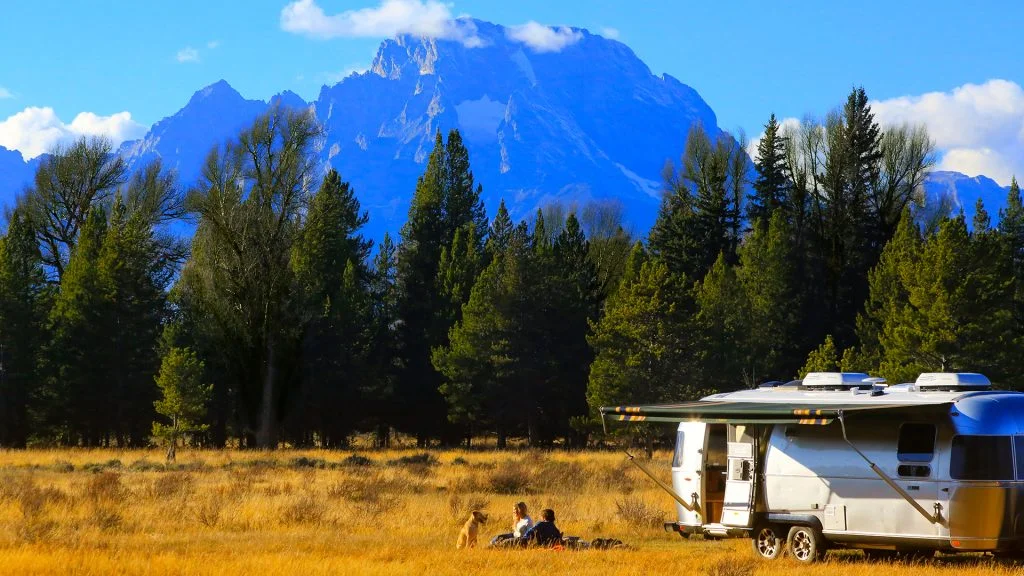 A silver camper sits in a grassy field near tall pine trees, with two people and a dog relaxing outside. Snowy mountains rise in the background under a bright blue sky with scattered clouds.