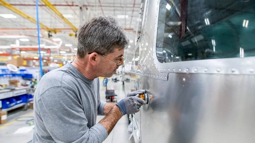 A man wearing glasses and gloves works on the exterior of a shiny metal aircraft inside a bright, spacious industrial facility.