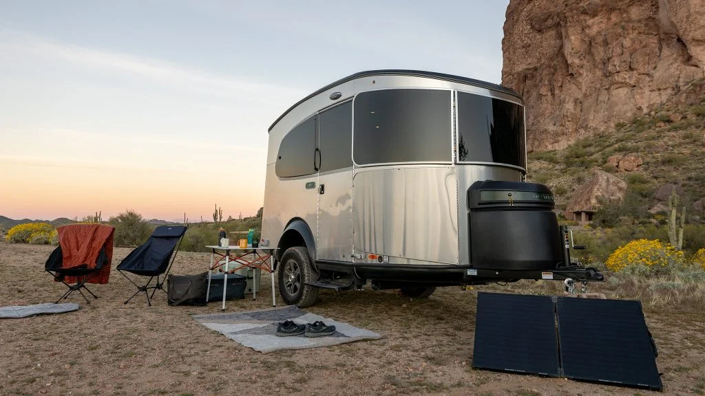 A sleek silver travel trailer parked in a desert campsite with chairs, a table, and solar panels, surrounded by rocks and desert plants.