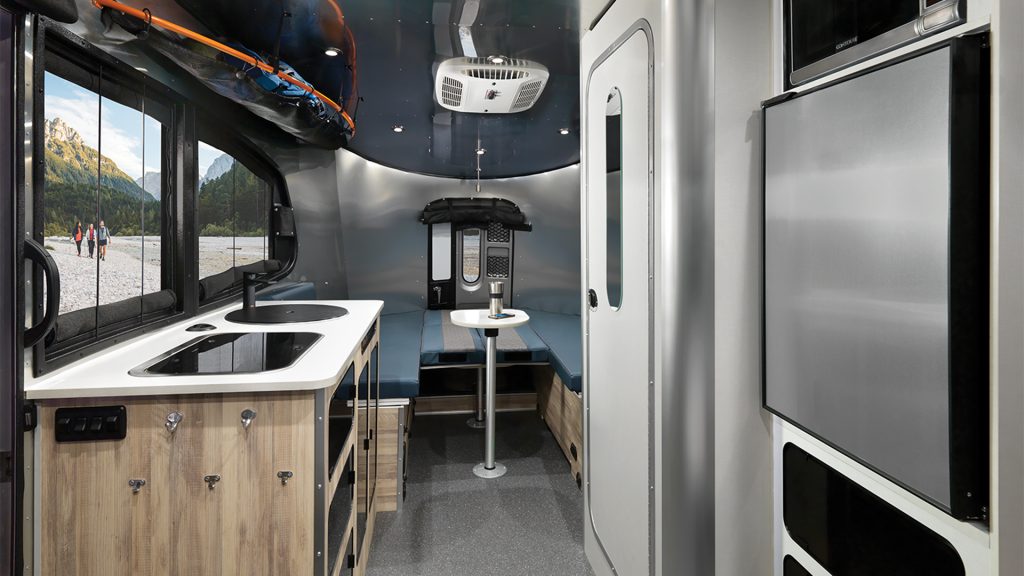 Interior of a modern camper van with a kitchenette, seating area, and large window showing a mountain view.