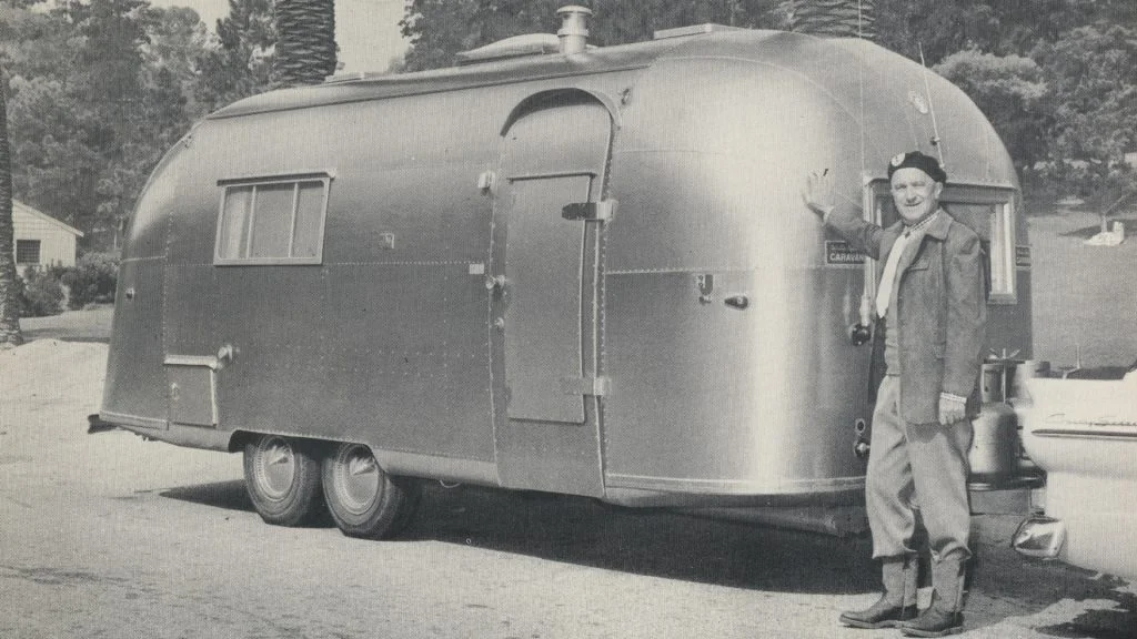 A man stands next to a vintage silver travel trailer in a black and white photograph, outdoors, with trees in the background.