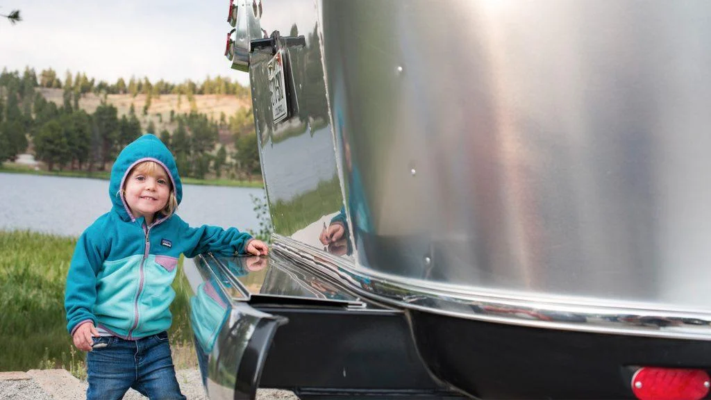 A young child in a turquoise hoodie stands beside a shiny silver trailer, smiling at the camera. A lake, green grass, and trees appear in the background under a cloudy sky.