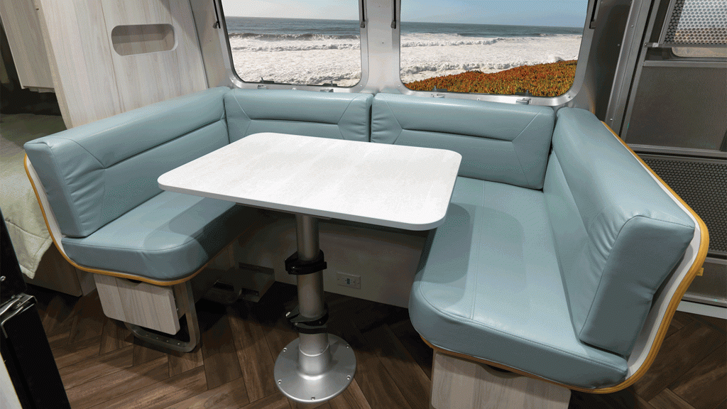 A modern RV interior with a U-shaped, light blue cushioned dinette and white table, positioned next to large windows showing a scenic ocean view.