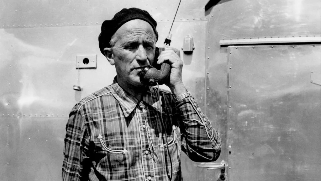 An older man wearing a plaid shirt and beret stands outside, holding a vintage telephone receiver to his ear in front of a metallic wall.