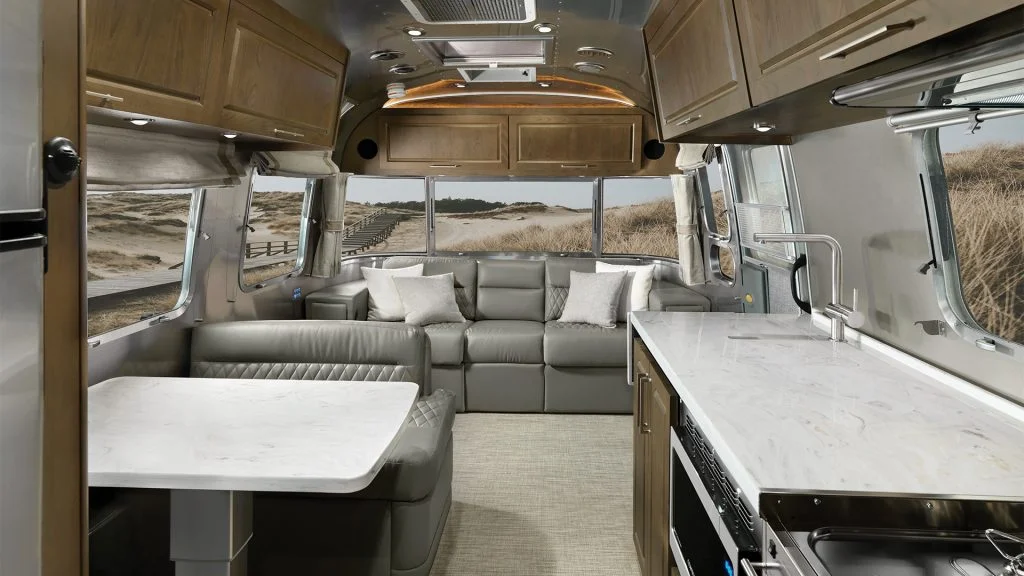 Interior of a modern RV with a gray sofa, white marble table, wooden cabinets, and large windows revealing a scenic view of sand dunes.