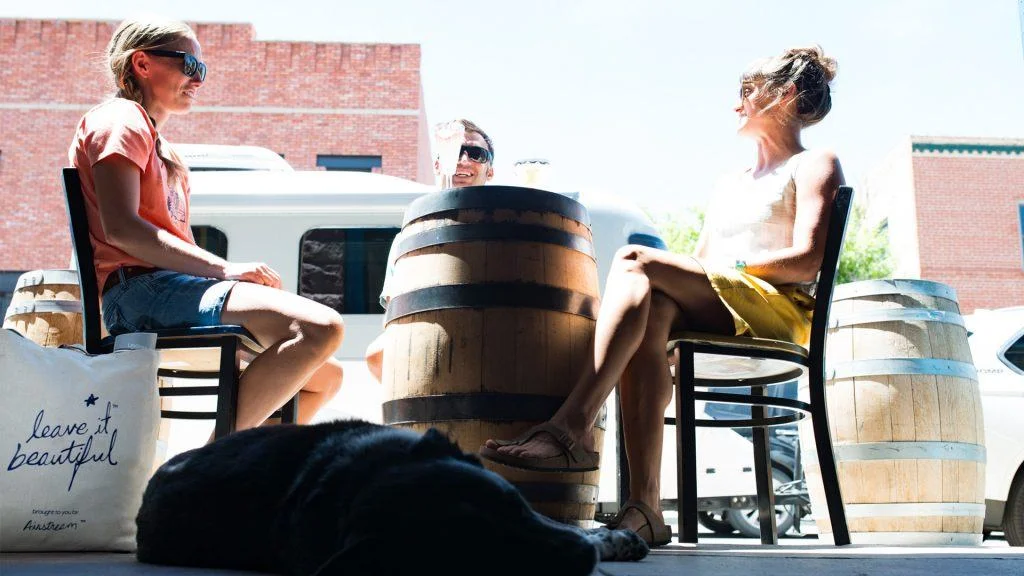 Three people sit around a barrel-shaped table outdoors, talking and laughing on a sunny day. A black dog lies on the ground in the foreground, and a tote bag nearby reads leave it beautiful. Brick buildings are in the background.