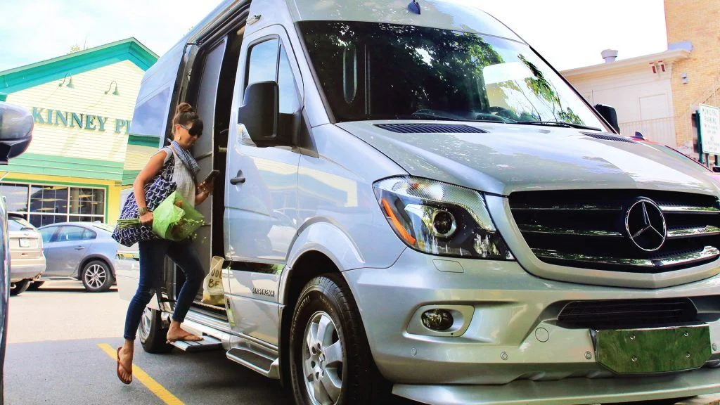 A woman holding groceries steps into a parked silver Mercedes-Benz Sprinter van in a parking lot, with shops and trees visible in the background.