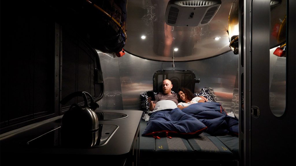 A couple relaxes on a bed in a cozy, dimly lit camper interior, with a compact kitchen area visible on the left.