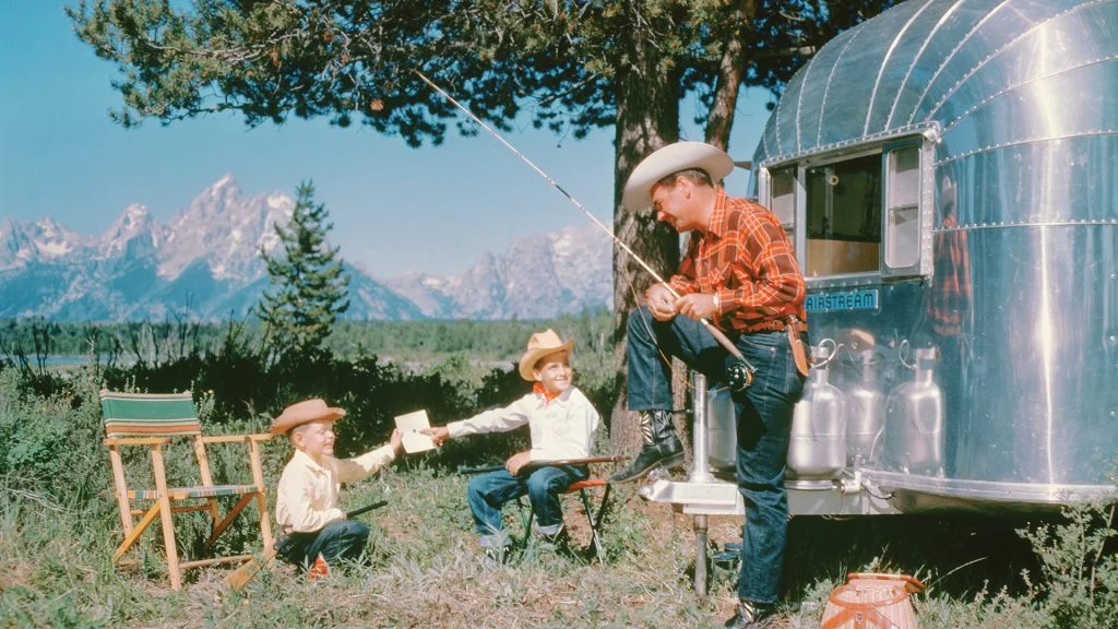 A man in a cowboy hat and plaid shirt sits by a shiny silver trailer, fishing. Two boys wearing cowboy hats sit nearby in front of mountains, one handing the other a piece of paper. Camping chairs and trees surround them.