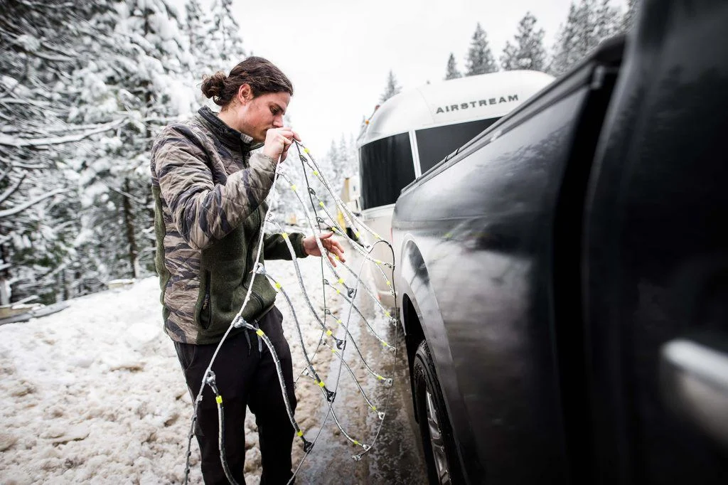 Person in a snow-covered forest attaching snow chains to a truck's tire, next to an Airstream trailer.