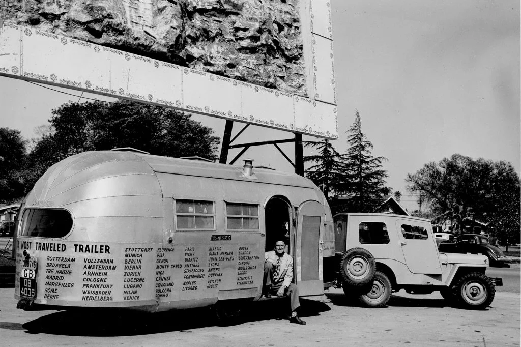 A vintage black-and-white photo of a man sitting in the doorway of a silver travel trailer, hitched to a classic jeep. Names of cities are written on the trailer. A large billboard and trees are visible in the background.