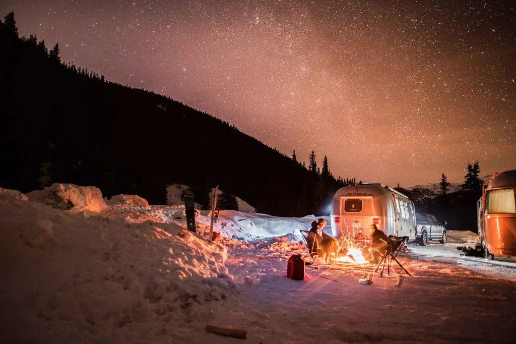 Camping scene with people around a campfire, surrounded by snow and parked trailers. A starry sky lights up the night.