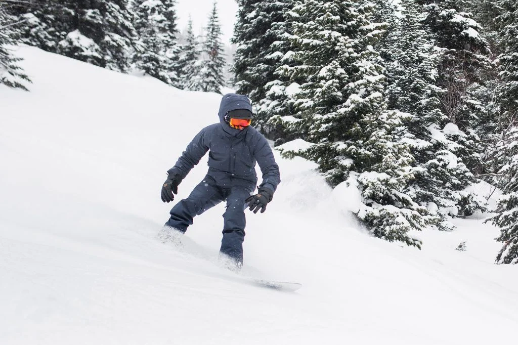 Snowboarder in dark gear gliding down a snowy slope, surrounded by snow-covered pine trees.