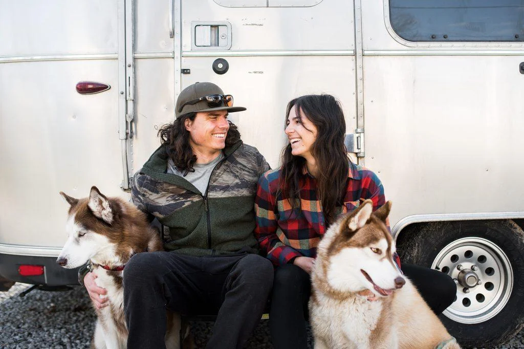 A couple sits smiling with two huskies in front of a silver trailer, enjoying a peaceful moment outdoors.