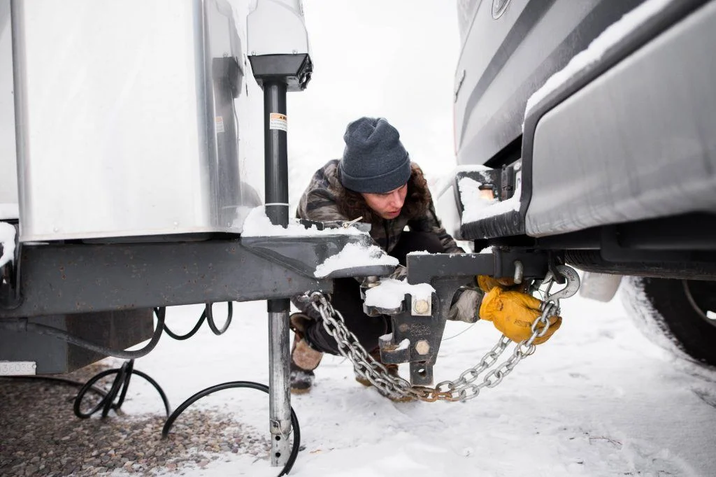 Person connecting a trailer hitch to a snowy vehicle, wearing a beanie and yellow gloves on a winter day.