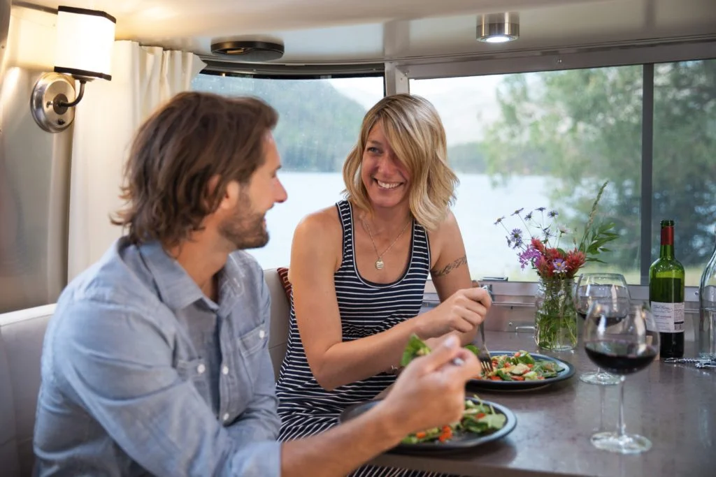 A man and woman enjoy a meal together inside an RV by a lake. They are smiling, with plates of salad, a vase of wildflowers, and a bottle and glasses of red wine on the table near a large window.