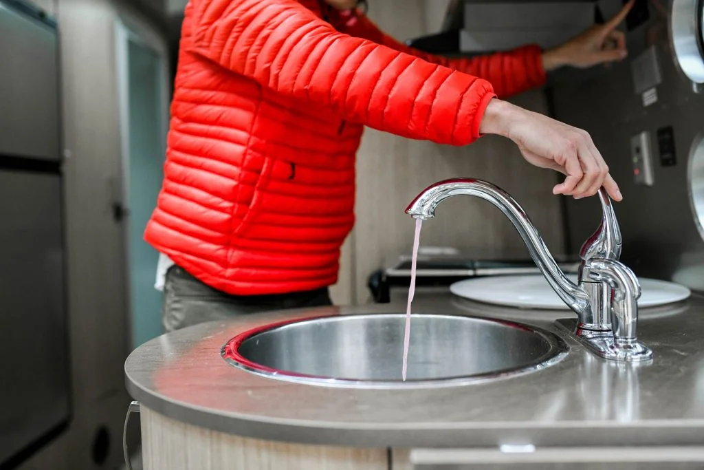 A person in a bright red jacket turns on a faucet in a modern RV kitchen, with water trickling into a round metal sink.