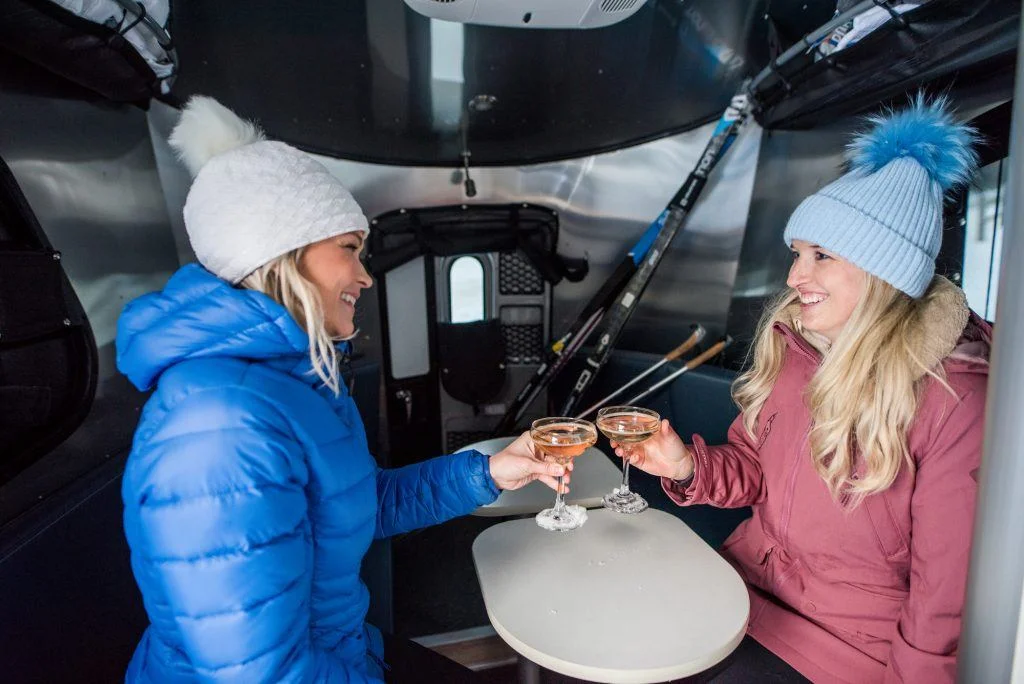 Two women in winter gear toast with drinks inside a cozy camper. Ski poles are visible in the background.