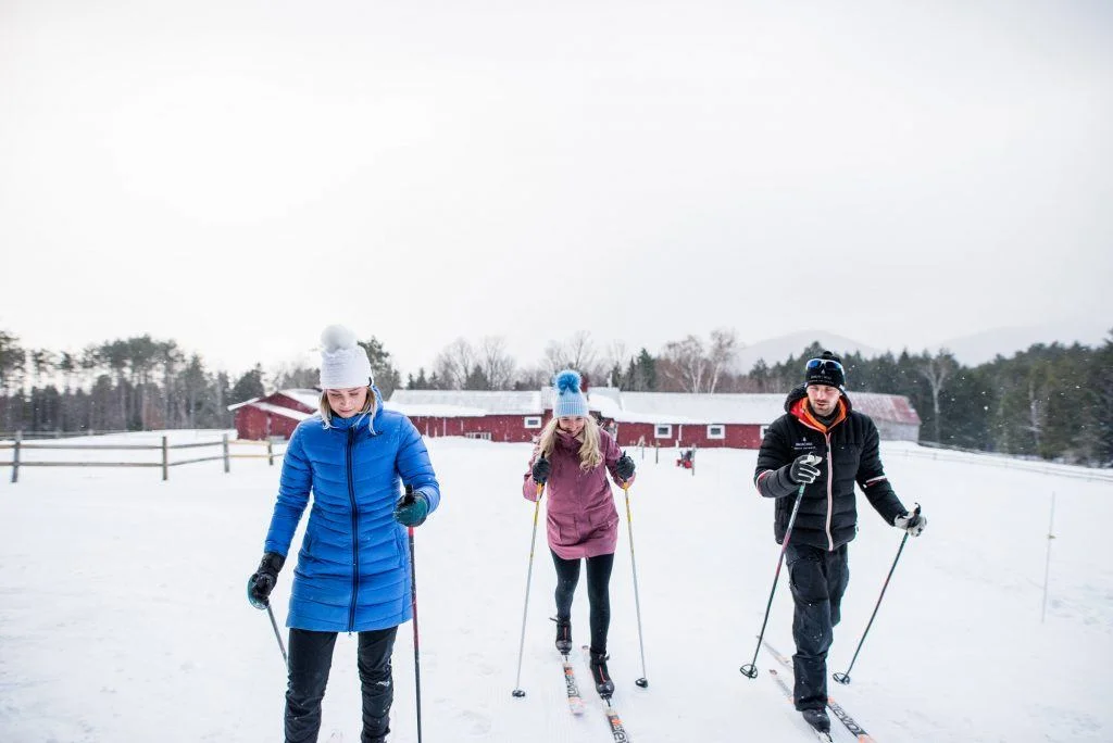 Three people cross-country skiing on a snowy trail by red buildings, with trees in the background, dressed in winter gear.