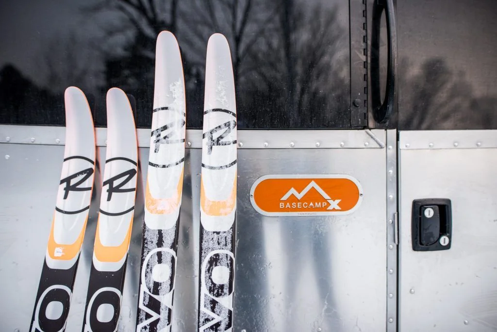 Four snow-dusted skis leaning against a metal trailer with an orange "Basecamp X" logo.