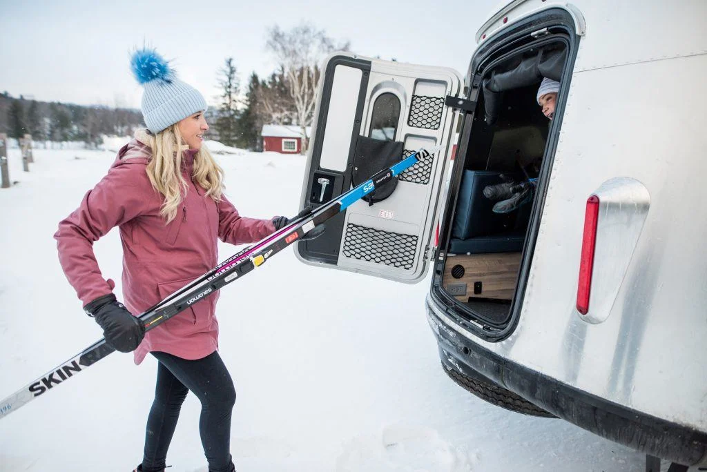 Woman in winter gear places ski poles in a trailer, with snow-covered landscape and trees in the background.
