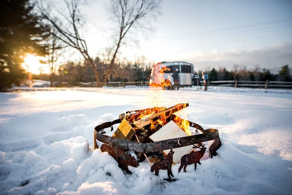 A fire pit with burning logs in a snowy landscape, featuring a metal ring with deer cutouts. A trailer and people are visible in the background.