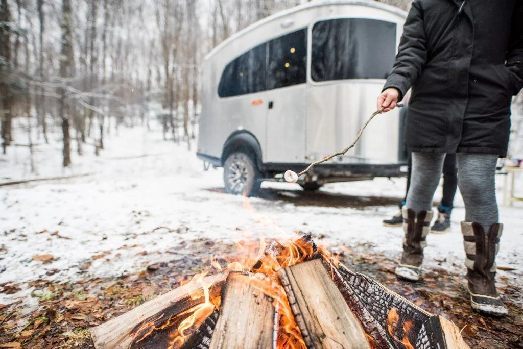 Person roasting marshmallows over a campfire near a silver trailer in a snowy forest setting.