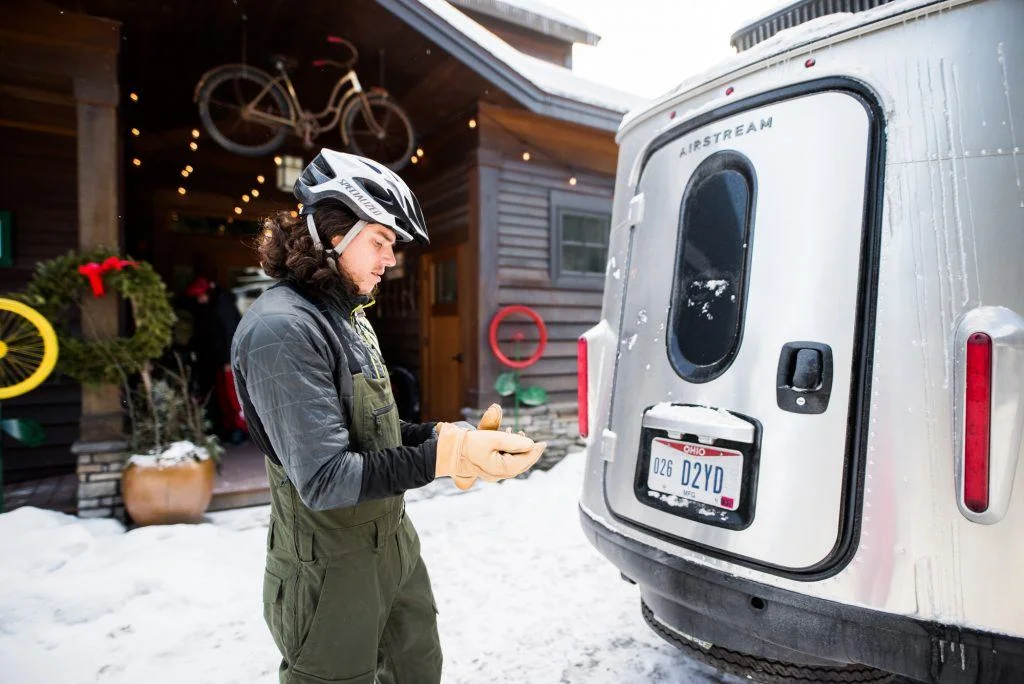 A person in winter gear and helmet stands near an Airstream trailer on snowy ground, next to a house decorated with wreaths and hanging bicycles.