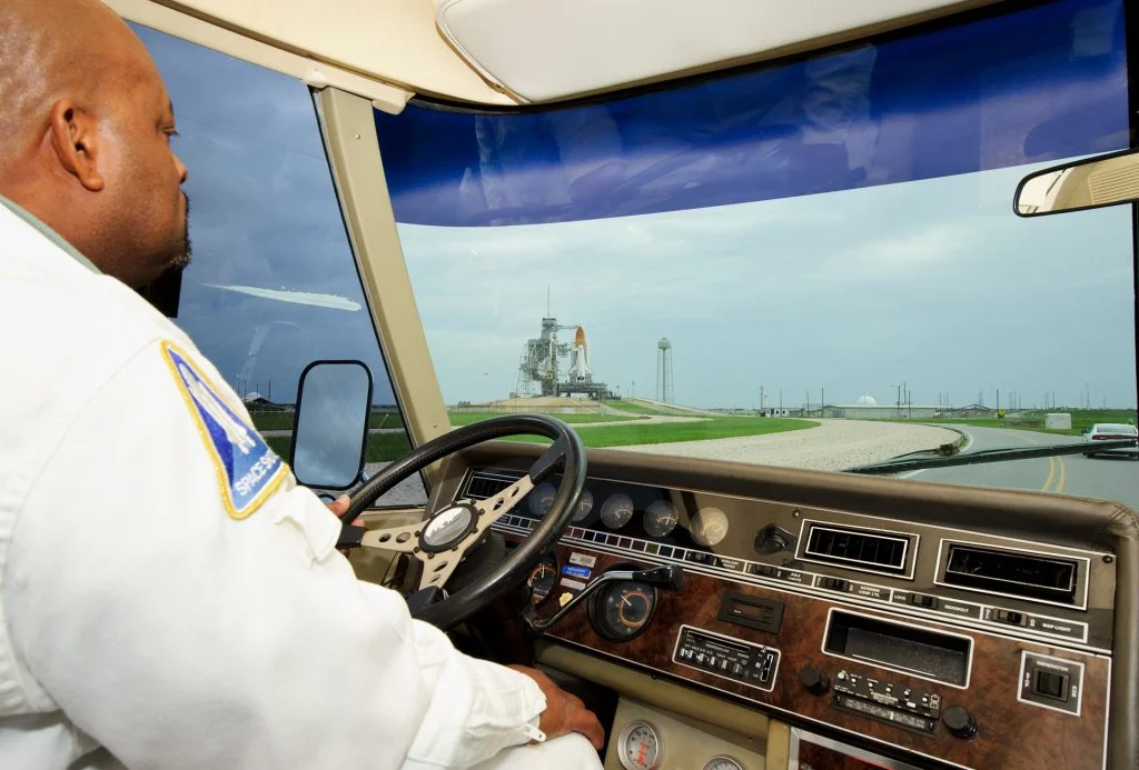 A person in a white uniform drives a vehicle toward a distant space shuttle on a launch pad under a cloudy sky, as seen through the windshield and dashboard.