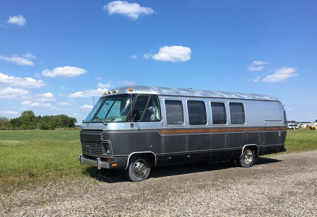 Silver vintage RV parked on a gravel path beside a green field, under a clear blue sky with scattered clouds.