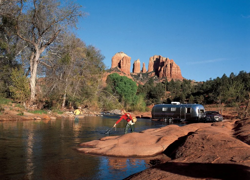 Two people fish in a shallow river near a silver Airstream trailer and vehicle, surrounded by red rocks and trees, with tall red rock formations in the background under a clear blue sky.