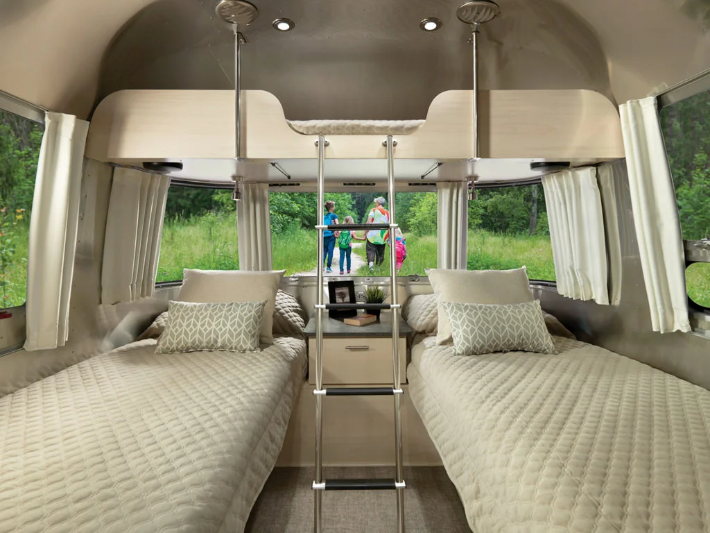 Interior of a modern camper with two twin beds, beige bedding, a small shelf with decor, a ladder to an overhead bunk, and large windows. Outside, a group of people stands on green grass in the background.