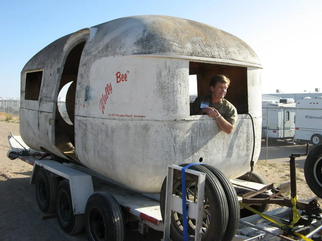 A man looks out from a worn, egg-shaped trailer labeled "Wild Goose," mounted on a multi-wheeled trailer, with vehicles in the background.